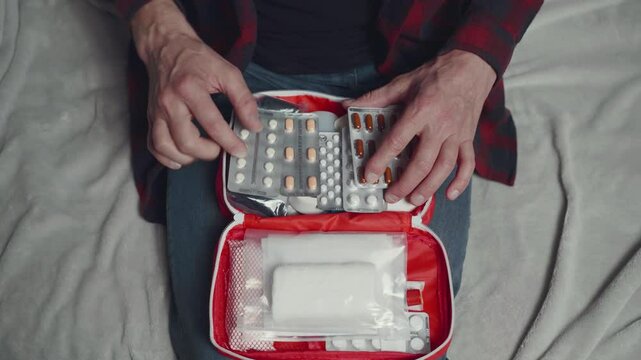 A closeup shows hands sorting medications in a red medical kit on a bed, highlighting the importance of organized health management and medication preparation for wellbeing