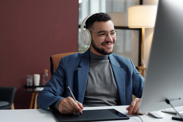 Portrait of smiling Middle Eastern man wearing headphones and using drawing tablet at workplace in office copy space
