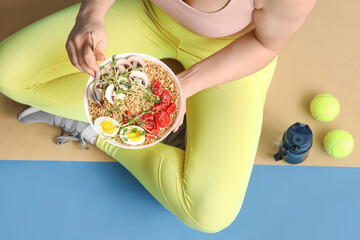 Sporty young woman sitting on yoga mat with bowl of nutritious oatmeal against blue background, top...