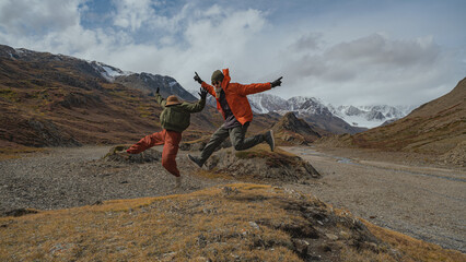 Two tourists in bright clothes make a jump in the mountains, and freeze in a funny pose. Behind them are the autumn mountains partially covered with snow.