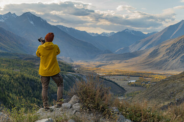 A man in bright clothes and a hat, photographing nature standing on a hill. The photographer takes pictures of a landscape gorge, overlooking a green forest, sky and mountains.