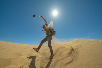 A traveler with trekking poles in his hands, with a backpack on his back, makes a jump from a sand dune, raising his arms and scattering sand with his feet.