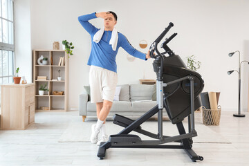 Tired young man near modern exercising bike at home