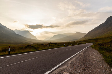 Naklejka premium Golden sunset over calm lakes and clear water reflections in The Trossachs and Loch Lomond. Long winding roads cross green hills and mountains in a peaceful, awe-inspiring natural landscape. Exploring