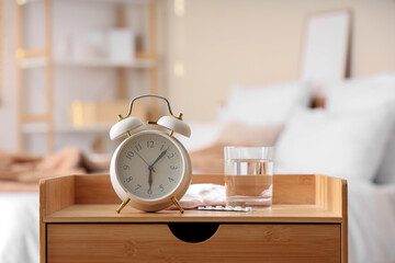 Alarm clock with glass of water and pills on bedside table in bedroom, closeup