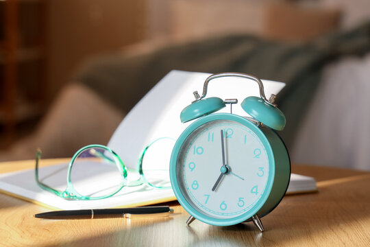 Alarm clock with notebook and eyeglasses on table in room, closeup