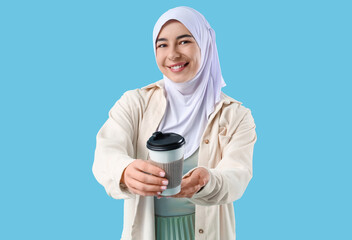Beautiful young Muslim woman in hijab with paper cup of hot coffee on blue background
