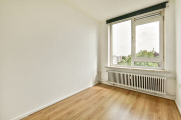 A spacious, bright room featuring a large window that offers a view of greenery outside. The walls are plain and the flooring is light wood, creating a minimalist vibe.