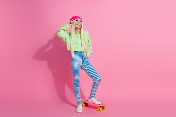 Stylish young blond woman posing playfully with a trendy outfit and pink skateboard against a vibrant pink studio background