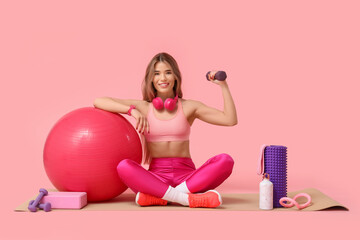 Sporty young woman with sports equipment sitting on yoga mat against pink background