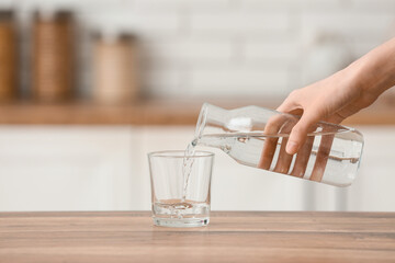 Woman pouring water from bottle into glass on wooden table in kitchen, closeup