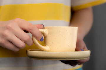 A woman in a yellow and white T-shirt with purple nail polish is holding a pale yellow cup and saucer with coffee. 
Close-up.
