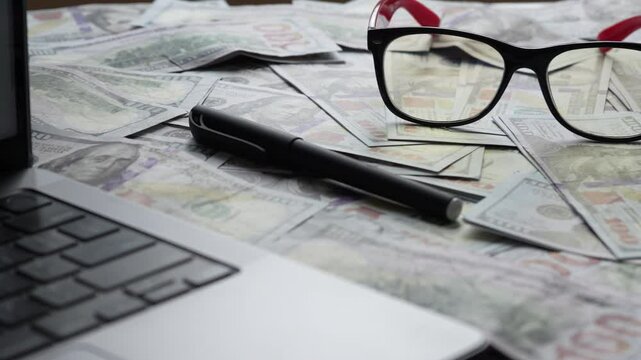 There is a laptop on the table, money is scattered nearby, hundred dollar bills, glasses and a wristwatch. Wiring to the right, close-up. Financial planning, saving, counting funds. 4k footage