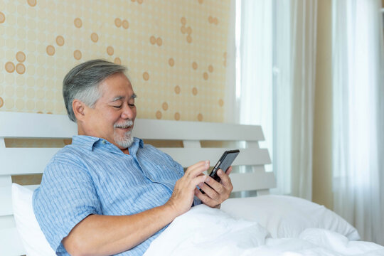 Happy Asian senior man using smartphone while relaxing on bed at home, elderly male enjoying online content with smiling face, retirement lifestyle and technology concept
