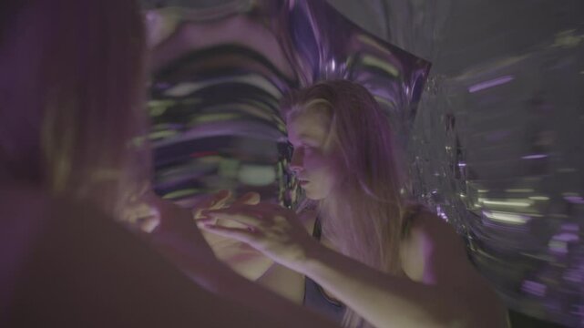 Young captivated woman dancing beside distortion mirror in multicolored illumination, looking at her altered reflection, giving artistic surreal performance
