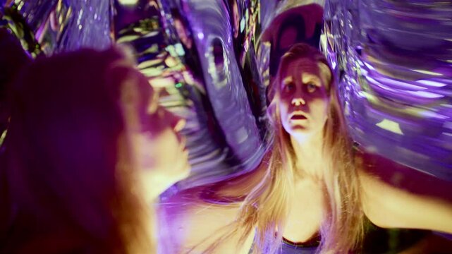 Young captivated woman gazing at fluid, transforming reflections in distorting mirror during dramatic performance under colorful neon lights