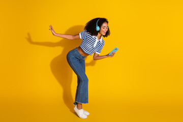 Young woman with headphones enjoying music and holding smartphone against yellow background...