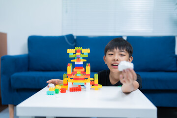 Fototapeta premium Asian boy is playing with colorful building blocks on a white table, showing a white block to the camera, with a blue sofa in the background, in a bright living room