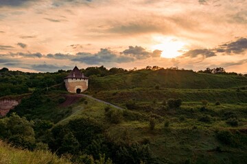 Watchtower of the architectural ensemble of the majestic medieval Khotyn fortress in Ukraine. Old stone tower with a pointed roof nestled on a grassy hill under a dramatic sky at sunset.