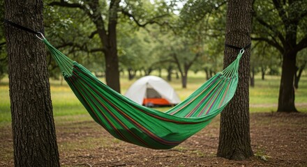 Peaceful camping scene featuring a green hammock strung between two trees with a tent set up in a serene natural environment surrounded by lush greenery