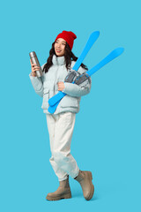 Young Asian woman in winter clothes with thermos and skis on blue background