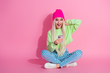 Portrait of a happy young stylish woman holding a smartphone while sitting against a vibrant pink background, showing excitement