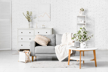 Sofa with dresser, coffee table and vase of blooming branches near white brick wall in living room