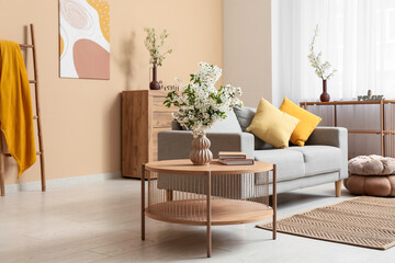 Sofa with dresser, coffee table and vases of white blooming branches near beige wall in living room