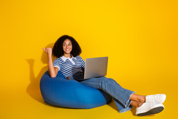 Smiling young woman with laptop relaxing on blue beanbag chair against vibrant yellow background