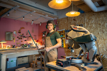 Experienced smiling carpenter stands ready at woodwork station holding measuring ruler with protective earmuffs. Workshop preparation, tool verification safety protocol check, precise measurements