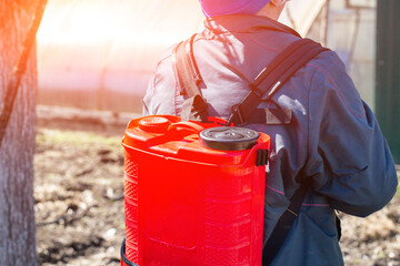 A man with a red electric sprayer treats trees and bushes in a garden on a summer cottage. Treatment of trees with copper sulfate in spring.