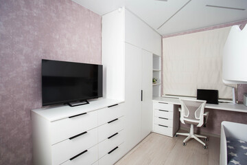 Modern workplace in the bedroom with white furniture, computer desk and chair. The TV is on a white chest of drawers. Bedroom interior.