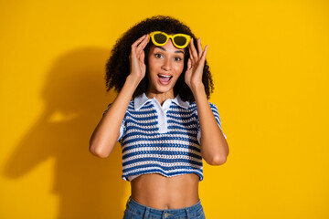 Cheerful young woman with curly hair wearing a vibrant blue top and yellow sunglasses against a bold yellow background