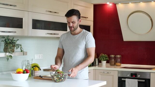 A man in a gray t-shirt is mixing a fresh salad in a modern kitchen. The counter is filled with colorful vegetables, fruits, and kitchen essentials, with a red wall in the background.