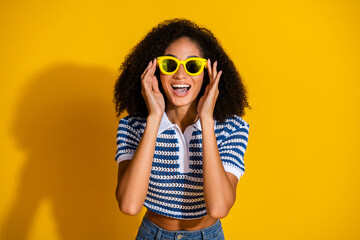 Cheerful young woman in stylish outfit with curly hair and sunglasses posing against a yellow background