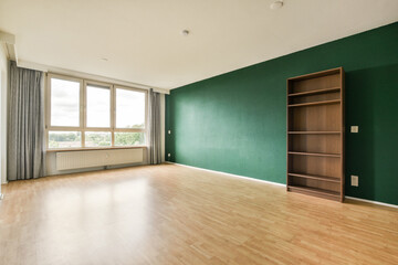 A spacious interior room featuring a striking green wall, large windows allowing natural light, and a wooden shelf emphasizing minimalism in design.