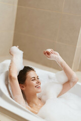 Joyful young woman relaxing in a bubble bath, enjoying a peaceful moment with arms raised, surrounded by foam and a soothing bathroom atmosphere.