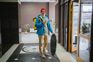 Courier wearing thermal backpack and holding skateboard is using smartphone app for navigation,...