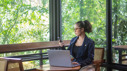 Asian businesswoman holding and staring at a smartphone with a serious expression while working on a laptop. Women using cell phones in cafe and remote work