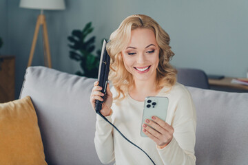 Young blonde woman using a flat iron while smiling at her phone in a cozy home living room with...