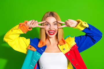 Happy young woman making playful peace sign gesture in colorful jacket with green background,...