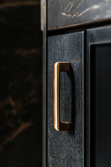 Close-up of modern bronze cabinet handle on dark wood panel in luxury kitchen setting, with soft lighting highlighting texture and design contrast.
