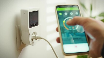 A person is using a smartphone to monitor energy usage via a smart plug device mounted on the wall. This activity occurs in a contemporary home, highlighting a focus on energy efficiency