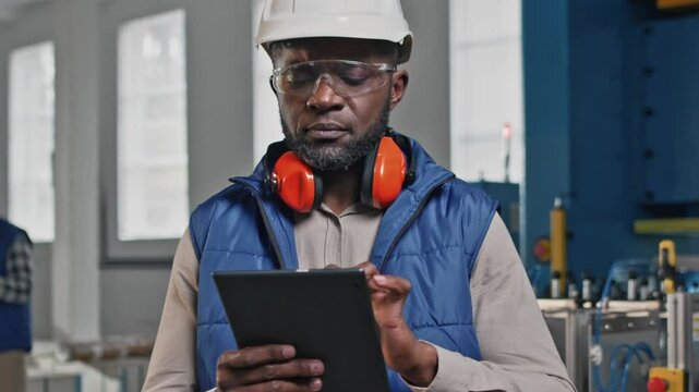 Serious African American engineer wearing helmet and safety glasses holding digital tablet in factory. Browsing technical documentation or entering production data while standing beside equipment.