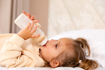Side view cute little toddler baby girl in bodysuit lying on bed with bottle of milk indoors....