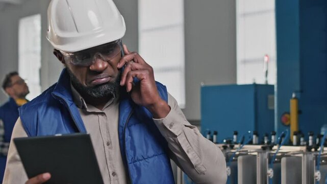 Concerned male technician wearing hard hat and transparent goggles talking on phone while holding tablet in production zone. Listening carefully while solving unexpected technical issue remotely.