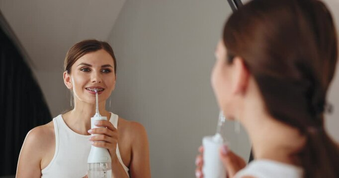 Young Caucasian woman with braces using dental irrigator in bathroom, smiling and looking in mirror for oral hygiene care.