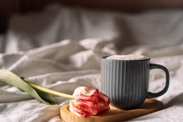 Coffee with foam on wooden stand next to tulip on white bed linen.