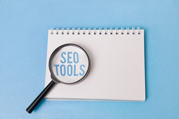 A magnifying glass with the words "SEO TOOLS" enlarged underneath rests on a blank notepad against a light blue background. Suggests exploring and researching resources for SEO