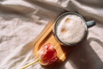 Coffee with foam on wooden stand next to tulip on white bed linen.